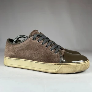 Bruna sneakers från Lanvin i mocka - Snygga bruna sneakers från Lanvin med ovandel i mocka och lackad tå i mörkbrunt. Skorna har grå snörning och en beige sula med retrovibbar. Perfekt för dig som gillar stilrena men unika detaljer. Klassisk rund tå och låg modell.