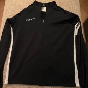 Svart Nike Dri-FIT träningströja - Svart långärmad träningströja från Nike med vita detaljer längs ärmarna och axeln. Tröjan har half zip framtill och är tillverkad i funktionsmaterialet Dri-FIT som andas. Perfekt för fotboll eller annan träning. Storlek L 147-158 