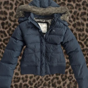 SÖKER Mörkblå pufferjacka Abercrombie - Snygg mörkblå pufferjacka från Abercrombie & Fitch med avtagbar huva och fuskpäls. Jackan har ribbade muddar, knappar och dragkedja framtill. Perfekt för kalla dagar och riktigt skön passform.