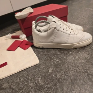 Valentino Rockstud vita sneakers - Säljer ett par vita Valentino Rockstud sneakers i skinn med ikoniska nitar på hälen och cool sula med camouflage-mönster undertill. Klassisk rund tå, snörning och diskret logga på plösen. Kommer med originalkartong, extra skosnören och dustbag.