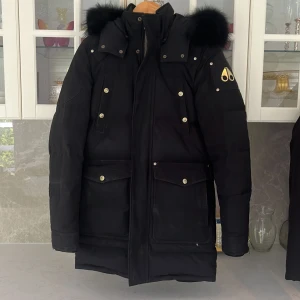 Svart dunjacka från Moose Knuckles - Säljer en svart dunjacka från Moose Knuckles med guldiga metalldetaljer och logga på ärmen. Jackan har stor huva med svart päls, flera fickor med tryckknappar och dragkedja framtill. Perfekt för kalla dagar och riktigt snygg streetstil. Speciell edition storlek S men funkar för storlek M. Kvitto finns