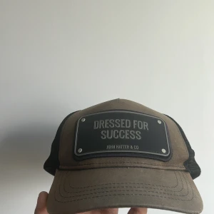 John Hatter & Co truckerkeps brun - Snygg truckerkeps från John Hatter & Co i brun och svart med mesh på baksidan och justerbar snapback. Framtill sitter en svart platta med texten 'Dressed for Success'. Perfekt för dig som vill sticka ut med en cool accessoar.