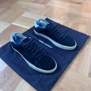 Saint Laurent SL/06 svarta  - Svarta Saint Laurent SL/06 sneakers i mocka med vit häl flärp. Snygg minimalistisk design med diskret logga på sidan. Skorna är i 9,5/10 skick, inprincip nya utan box. Skorna är i storlek it 40 men passar 40-41,5. Riktigt sköna skor som går lätt under radarn. Hör av dig vid minsta intresse. 