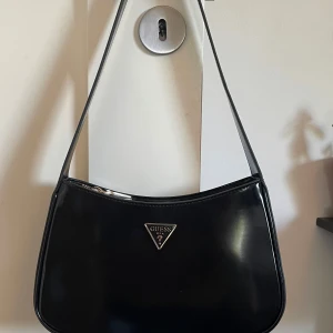 Guess Väska - Snygg svart axelväska från Guess med stilren, glansig finish och klassisk trekantslogga framtill. Väskan har ett huvudfack med dragkedja och innerficka, perfekt för det viktigaste. Tillverkad i syntetmaterial och har en enkel, modern form. Aldrig använd, som ny. Nypris: 1500kr