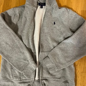 Grå hoodie från Polo Ralph Lauren - Mysig grå hoodie från Polo Ralph Lauren med dragkedja och broderad logga på bröstet. Fodrad med mjuk fleece på insidan och har långa ärmar samt praktiska fickor framtill. Perfekt för kyliga dagar och enkel att matcha med allt. Barn storlek