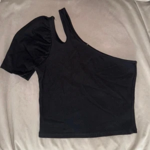 Svart oneshoulder topp med puffärm - Säljer en svart oneshoulder topp med en kort puffärm på ena sidan och axelbandslös på andra. Toppen är croppad och har en stilren, asymmetrisk design som ger en cool vibe. Perfekt att styla med höga jeans eller kjol. Toppen är i mycket fint skick!