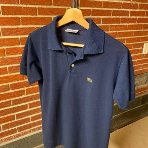 Mörkblå pikétröja från Lacoste - Klassisk mörkblå pikétröja från Lacoste med korta ärmar och ikoniska krokodilloggan broderad på bröstet. Tröjan har krage och två knappar framtill. Tillverkad i bomull för en skön och luftig känsla.