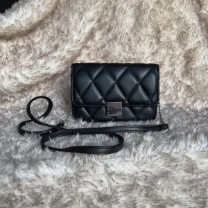 Svart quiltad axelväska med kedja - Snygg svart axelväska i quiltad design. Väskan har en lång axelrem i skinnimitation och silverfärgad kedjedetalj. Stängs med ett fyrkantigt spänne framtill. Tom kan väskan se lite ojämn ut man annars fin. Bredd 19 cm, Längd 14 cm. Längd med rem ca 70 cm.