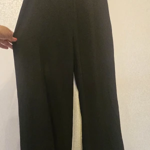 Svarta vida kostymbyxor - Säljer ett par svarta kostymbyxor med hög midja och vida ben. Byxorna har en klassisk look och är tillverkade i ett mjukt tyg som faller snyggt. Perfekta för dig som gillar en stilren och trendig vibe.