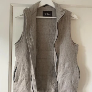 Beige quiltad väst från Oscar Jacobson - Snygg beige quiltad väst från Oscar Jacobson med hög krage och två fickor med dragkedja. Västen har en stilren design och är tillverkad i ett mjukt material som ger en chill och clean look. Perfekt att slänga över en hoodie eller tröja för extra värme.      Använd 1-2 gånger . Nypris 1299:- En liten xs Med måtten  ca 82cm i midja Ca 76cm bröst Och ca 56 cm ifrån linningen upp till halsen
