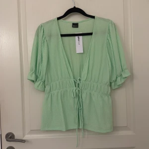 Mintgrön blus från Glamorous - Superfin mintgrön blus från Glamorous med v-ringning, knytband i midjan och puffiga korta ärmar. Blusen har en luftig och lätt struktur, perfekt för vår och sommar.