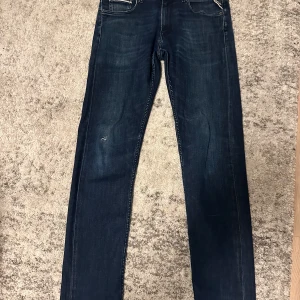 Replay Grover jeans mörkblå herr - Säljer ett par Replay Grover jeans i mörkblå tvätt med klassisk femficksdesign. Jeansen har raka ben, diskret slitning på låret och snygga detaljer vid fickorna. Perfekta för dig som gillar en tidlös och stilren look. OBS: litet hål/slit spår på vänster benet (se bild 4).