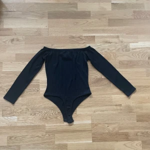 Svart ribbad offshoulder body - Snygg svart body med ribbad struktur och offshoulder-design. Långärmad modell som sitter tight och framhäver figuren. Perfekt att styla med jeans eller kjol för en trendig look.