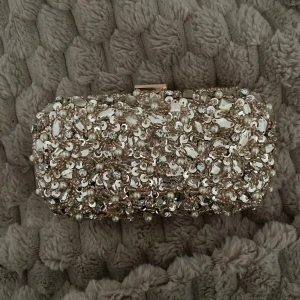 Glittrig kuvertväska - Säljer en superfin kuvertväska från Mascara i champagne med massor av pärlor, paljetter och glittriga stenar på framsidan. Baksidan är helt glittrig och insidan är fodrad i ljus satin. Perfekt när du vill sticka ut och addera lite extra bling till din look. Band följer med! Originalpris var över 500kr. 