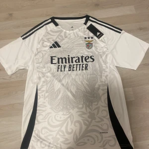 Vit Benfica fotbollströja Adidas L - Snygg Benfica fotbollströja från Adidas i storlek L. Tröjan är vit med svarta detaljer, klubbmärke och sponsortryck framtill. Unikt mönster över hela tröjan och klassiska Adidas-ränder på axlarna. Tillverkad i lätt och ventilerande material.