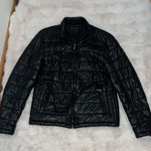 Svart quiltad pufferjacka Zara Man XL - Svart quiltad pufferjacka från Zara Man i storlek XL. Jackan har en stilren look med dragkedja framtill, två fickor med dragkedja och lätt glansig yta. Tillverkad i syntetmaterial med vaddering, perfekt för kyligare dagar.