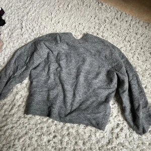 Grå stickad tröja med rund hals - Mysig grå stickad tröja med rund halsringning och långa ärmar. Tröjan har en enkel och stilren design med ribbade muddar vid ärmslut och nederkant. Perfekt för lager-på-lager under kyliga dagar.