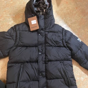 Svart pufferjacka från Burberry - Säljer en svart pufferjacka från Burberry med huva och dragkedja. Jackan har klassiskt rutigt foder i huvan och två fickor med dragkedja framtill. Perfekt för kalla dagar och riktigt snygg streetstil.