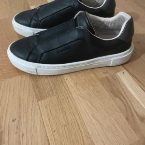 Svarta sneakers från Calvin Klein - Stilrena svarta sneakers från Calvin Klein i skinn med vit sula och diskret logga på sidan. Skorna har en minimalistisk design utan snörning och en mjuk, ljus insida för extra komfort. Perfekta för dig som gillar en clean och modern look.