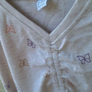 Beige ribbad långärmad topp med fjärilar - Superfin beige ribbad långärmad topp med v-ringning och rynkdetalj framtill. Toppen har små fjärilar i lila, orange och beige tryck över hela tyget. Perfekt för dig som gillar söta detaljer och mjuka material.