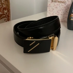 Svart skärp med guldfärgat spänne - Snyggt svart bälte i skinn med ett stilrent, rektangulärt spänne i guldfärg och svarta detaljer. Spännet har två diagonala guldfärgade linjer som ger en modern vibe. Bältet har en slät yta och justerbar längd med smart låsmekanism.