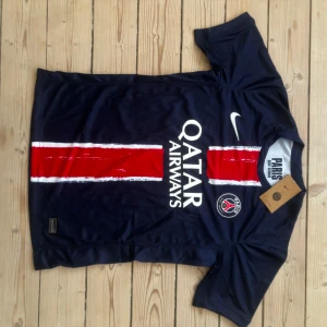 PSG Nike matchtröja blå röd herr S - Paris Saint-Germain fotbollströja från Nike i Dri-FIT-material. Djupblå med röd vertikal rand och vita detaljer, PSG-logga och Qatar Airways-tryck på bröstet. Kortärmad modell, perfekt för match eller träning.