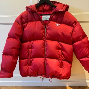 Röd pufferjacka från Prada, strl 38 - Knappt använd-som ny! Riktigt fet röd pufferjacka från Prada med ikonisk logga på bröstet. Jackan är i Re-Nylon, har dragkedja framtill, huva och dragsko nertill. Oversized vibe och perfekt för kalla dagar. Snygg och statement, passar dig som vill sticka ut.