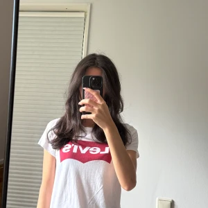 Vit Levi's t-shirt med röd logga XS - Vit t-shirt från Levi's i mjuk bomull med klassisk röd logga framtill. Modellen är kortärmad och har en rund halsringning. Perfekt basic-plagg med snygg streetkänsla och enkel passform.