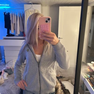 Grå hoodie från Lyle & Scott - Grå hoodie från Lyle & Scott med dragkedja och huva. Klassisk gul logga på bröstet och två fickor framtill. Mjuk bomullskänsla och långärmad modell, perfekt för chill dagar.