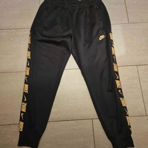 Svarta Nike mjukisbyxor stl M - Svarta mjukisbyxor från Nike med guldiga Nike-loggor längs sidorna och en liten logga framtill. Byxorna har resår i midjan och vid bensluten, samt sidofickor. Perfekta för en sportig och avslappnad stil. Nypris ca 800 kr.