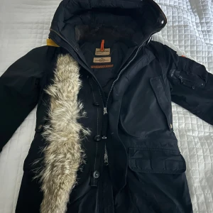 Svart Parajumpers parkas med pälsdetalj - Säljer en svart Parajumpers parkas med huva och avtagbar pälsdetalj. Jackan har flera fickor, dragkedja och knappar framtill samt ikonisk gul rem på ryggen. Insidan är quiltad för extra värme och är perfekt för kalla dagar.