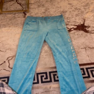 Blå velourbyxor med strassdetaljer - Säljer ett par ljusblå velourbyxor med raka ben och stora fickor fram och bak. Byxorna har elastisk midja och glittrande strassdetaljer längs ena benet. Perfekta för dig som gillar att sticka ut med en mjuk och skön stil. Har complete så 500kr för båda nya skick 