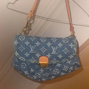Väska  - Säljer en snygg axelväska i blått jeanstyg med ikoniskt monogrammönster. Väskan har guldfärgade metalldetaljer, läderrem i beige och orange samt ett klassiskt spänne framtill. Perfekt för dig som vill sticka ut med en trendig designerbag.