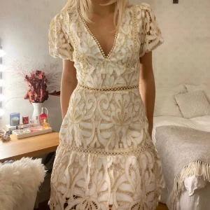 Vit spetsklänning  - Superfin vit kortklänning med v-ringning, puffärmar och broderade spetsdetaljer i beige. Klänningen har ett vackert mönster, markerad midja och volangkant nertill. Perfekt för sommarens alla tillfällen!