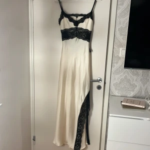Lång satinklänning med spetsdetaljer - Supersnygg lång klänning i ljus satin från Gina tricot. med tunna axelband och svarta spetsdetaljer vid ryggen, midja och längs slitsen. Klänningen har en öppen rygg och figurnära passform som ger en elegant siluett. Perfekt för dig som vill sticka ut.