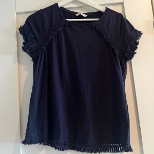 Mörkblå topp med fransdetaljer Ellos - Snygg mörkblå topp från Ellos i bomull med korta ärmar. Toppen har dekorativa fransar och bollar längs ärmar och nederkant
