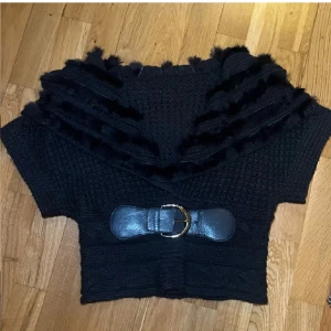 Mörkblå stickad kofta med bälte och pälsdetaljer - Snygg svart stickad kofta med korta ärmar och ett brett bälte i midjan. Koftan har en unik krage med pälsdetaljer. Lite slitningar i bältet men inget som syns om man inte kollar väldigt noggrannt! Materialet består av 35% ull, 25% bommul och 40% akryl, perfek till kyligare dagar. Färgen är väldigt mörkblå, nästintill svart. Storlek S, normal i storlek och bältet är justerbart.