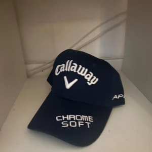 Mörkblå Callaway keps med brodyr - Snygg mörkblå keps från Callaway med vit broderad logga framtill och texten 'CHROME SOFT'. På sidan står 'APEX' och baktill finns Odyssey-loggan i vitt och rött. Justerbar rem bak för perfekt passform. Perfekt för golf eller chill. Ny pris 329kr mitt pris 199kr, kepsen är helt ny, Hör av dig om du undrar något