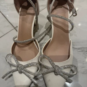 Vita pumps med glittriga rosetter - Snygga vita pumps med spetsig tå och glittriga remmar som går runt vristen. Framtill sitter stora rosetter i strass som ger extra bling. Perfekta för dig som vill sticka ut med eleganta detaljer och en feminin look.