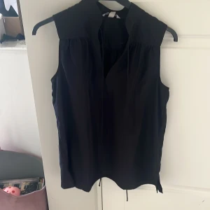 Svart ärmlös blus från H&M - Svart ärmlös blus från H&M med v-ringning och lätt rynkade axlar. Blusen har en lös passform och är tillverkad i ett mjukt, tunt tyg som känns skönt mot huden. Perfekt för en stilren och enkel look.