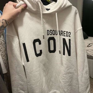 Vit hoodie från Dsquared2 ICON - Säljer en vit hoodie från Dsquared2 med stor svart 'ICON'-text framtill. Tröjan har huva med snörning och långa ärmar. Oversized fit och tillverkad i mjuk bomull. Perfekt statement-plagg för dig som gillar streetstyle.