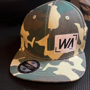 Warp keps med camouflage-mönster - Snygg keps från Warp med klassiskt camouflage-mönster i grönt, beige och brunt. Kepsen har rak skärm, justerbar snapback och en vit patch med svart logga framtill. Perfekt för dig som gillar streetwear och vill sticka ut med en cool accessoar.