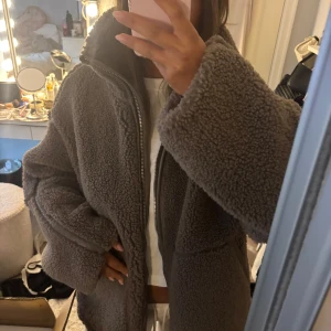 Brun oversized teddyjacka med dragkedja - Mysig brun teddyjacka med oversized passform och stora ärmar. Jackan har hög krage och stängs med dragkedja framtill. Perfekt för kalla dagar när du vill vara både varm och stylish.