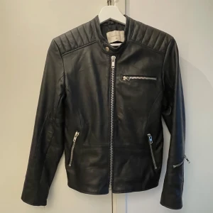 Snygg  skinnjacka – Edgy & Stilren look! - Snygg svart skinnjacka med dragkedjor, quiltade axlar , fodrade och figurnära passform. Hög krage med tryckknapp – perfekt för en cool look!