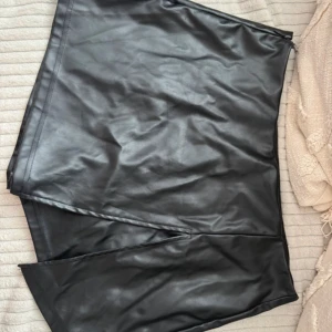 Svart kort kjol i skinnimitation - Snygg svart kort kjol i skinnimitation med slits framtill. Kjolen har en stilren design och dragkedja i sidan för enkel på- och avtagning. Perfekt för dig som gillar en edgy och trendig look.