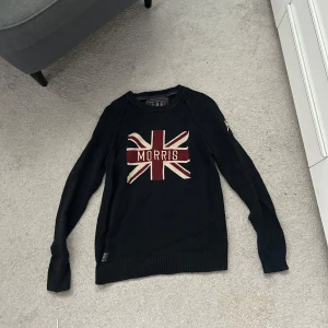 Svart stickad tröja Morris Union Jack - Svart stickad tröja från Morris med Union Jack-flagga och texten 'Morris' på bröstet. Tröjan har rund halsringning, ribbade muddar och långa ärmar. Perfekt för dig som gillar klassisk brittisk stil.