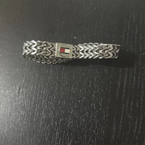 Tommy Hilfiger silverfärgat armband - Snyggt armband från Tommy Hilfiger i flätad silverfärgad metall. Låset har märkets klassiska logga i rött, vitt och blått. Perfekt accessoar för dig som gillar stilrena detaljer med en sportig touch.Ny pris 900 kr