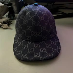 Gucci keps, S 56cm - Snygg blå keps från Gucci med det klassiska GG-mönstret över hela kepsen. Kepsen har en justerbar rem baktill. Materialet är jeansliknande och insidan är ljus med märkning. Perfekt för dig som vill ha en lyxig streetwear-look. Priset går och diskutera vid snabb affär. 