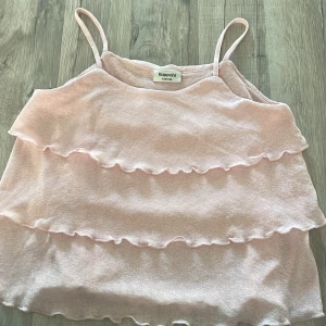 Ljusrosa volanglinne från Kappahl - Supersöt ljusrosa linnetopp från Kappahl med tunna axelband och flera volanger framtill. Toppen är ärmlös och har en luftig känsla, perfekt för varma dagar. Materialet är mjukt och lätt, vilket gör den extra skön att bära.