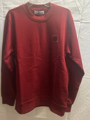 Vinröd sweatshirt från Stone Island - Säljer en vinröd sweatshirt från Stone Island med klassisk rund hals och broderad logotyp på bröstet. Tröjan är i mjuk bomull och har långa ärmar samt ribbade muddar vid ärmslut och nederkant. Perfekt för dig som gillar stilrena och tidlösa plagg.
